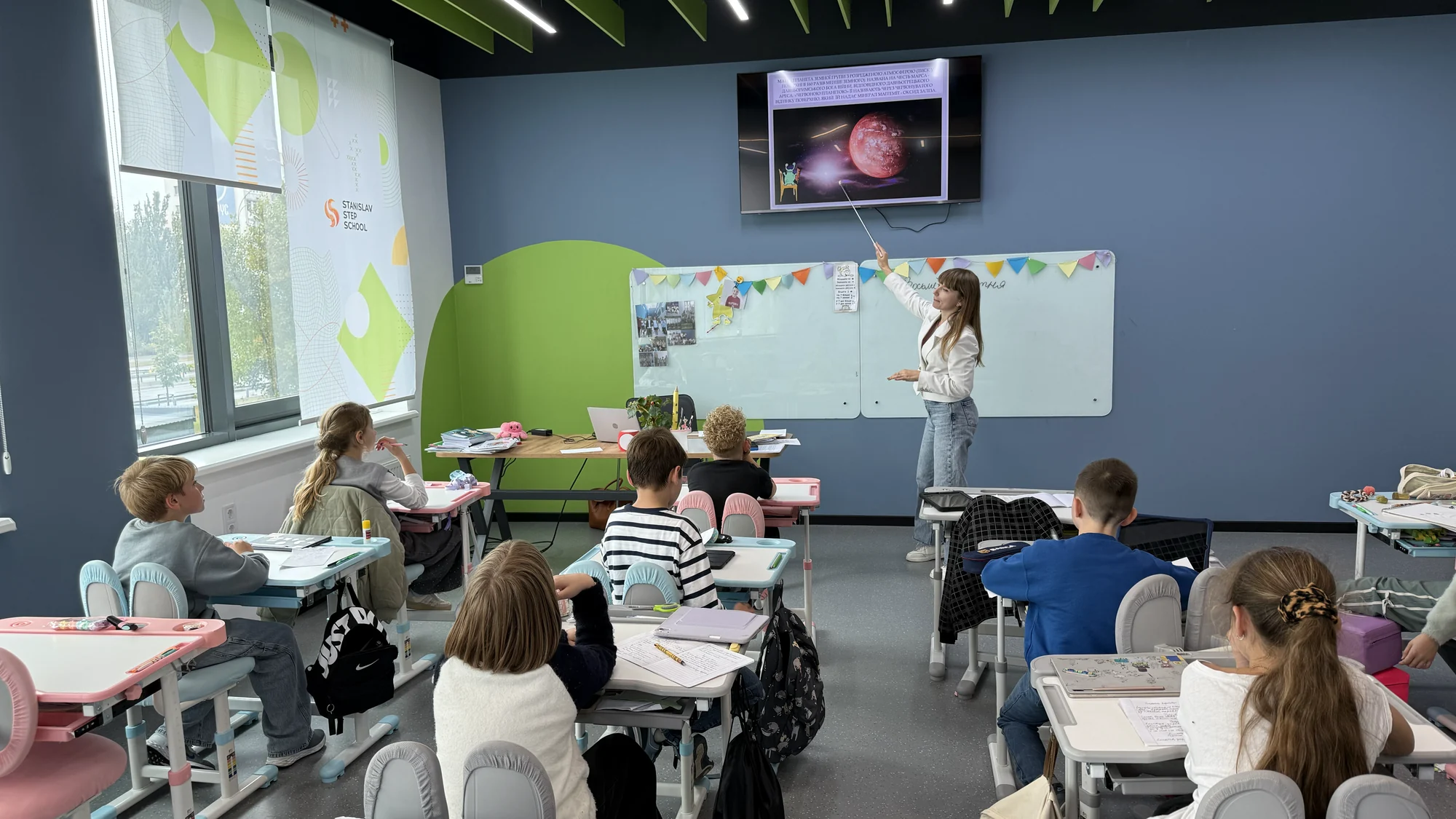 Урок у класі початкової школи: діти за кольоровими партами, вчителька біля екрана